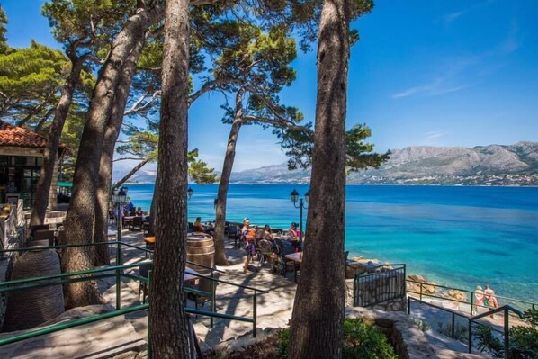 A seaside terrace with outdoor seating, overlooking a clear blue sea and distant mountains.