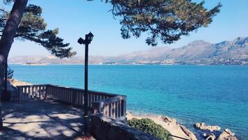A coastal walkway with a stone railing, a lamppost, and a view of a calm sea and distant mountains.