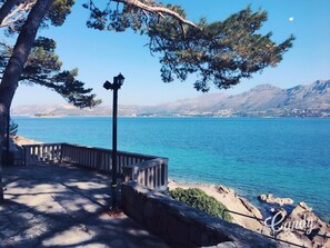 A coastal walkway with a stone railing, a lamppost, and a view of a calm sea and distant mountains.