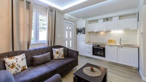 A modern living room with a sofa and a coffee table, a kitchen with white cabinets and appliances, and a window with curtains. - Vacation house Cavtat (Konavle)