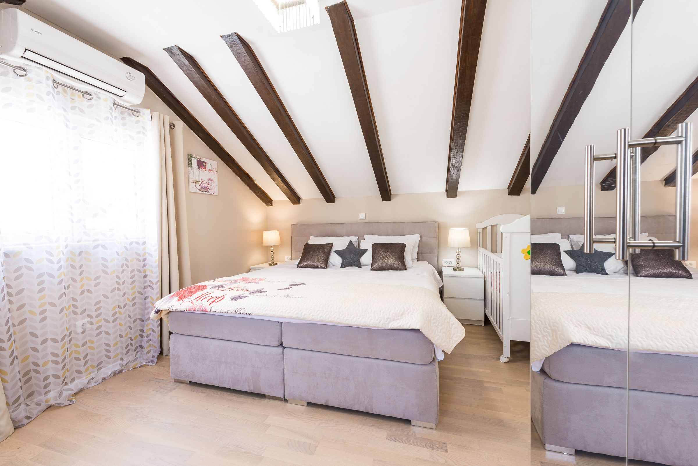 A bedroom with a large bed, a smaller bed, a nightstand, and a crib. There is a window with curtains, a ceiling fan, and wooden beams.