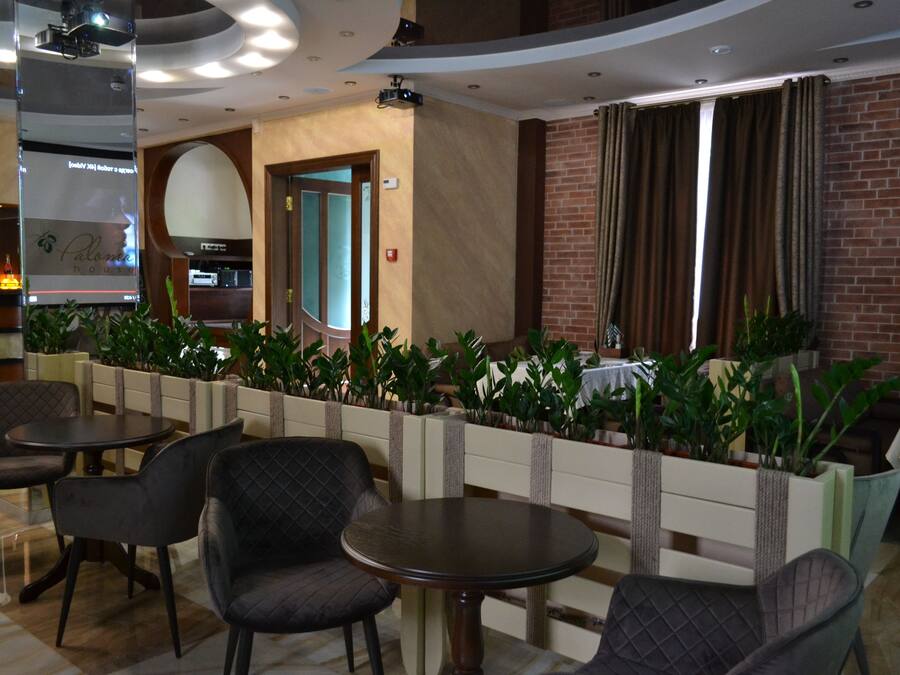 A modern indoor dining area with round tables and cushioned chairs, a brick wall, and a plant barrier.