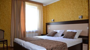 A double bed with a brown bedspread and two pillows, a wooden nightstand, a chair, and a window with curtains. - Paloma House (Kharkiv)