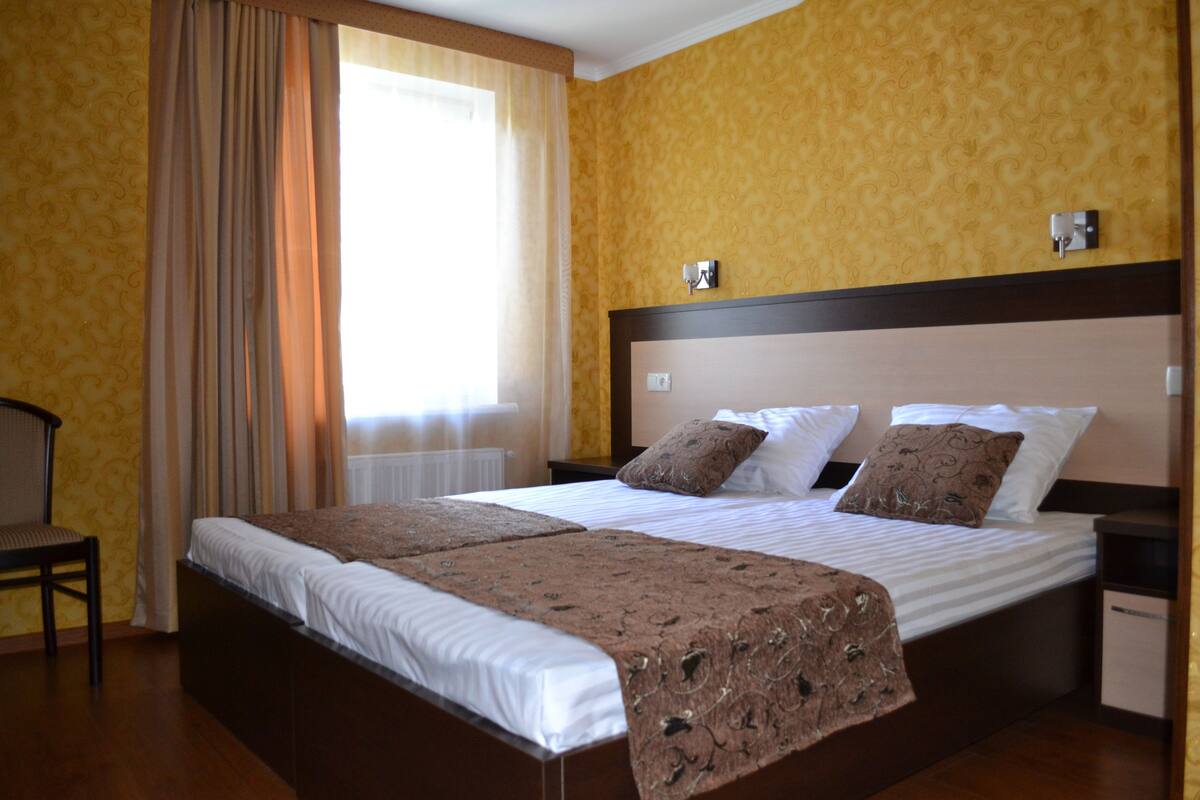 A double bed with a brown bedspread and two pillows, a wooden nightstand, a chair, and a window with curtains.