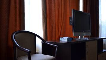 A hotel room with a desk, a television, a chair, and a window with curtains.