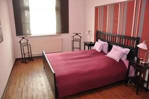 Un dormitorio con suelo de madera, una cama con colcha roja, un radiador, una ventana con persianas y una pared a rayas.