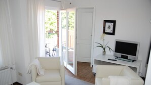 Ein Wohnzimmer mit einem weißen Sofa, einem weißen Sessel, einem Fernseher auf einem Standfuß und einer Glasschiebetür, die zu einem Außenbereich mit einem Patio-Tisch und -stühlen führt.