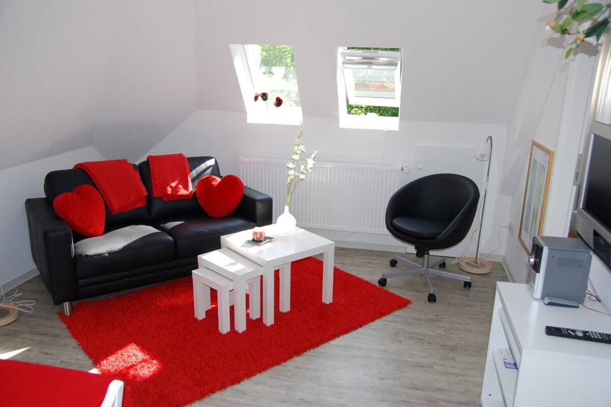 A modern living room with a black sofa, red cushions, a white coffee table, and a red rug. There is a black office chair, a radiator, and a window with a view of greenery.