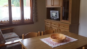 A dining area with a wooden table, chairs, a television on a cabinet, and a window with curtains. - Casa Rosina 6 (Val di Zoldo)