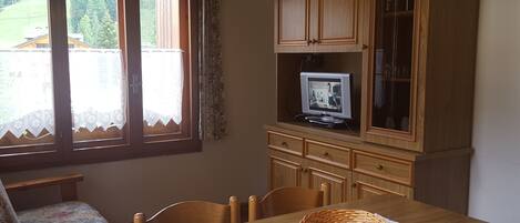 A dining area with a wooden table, chairs, a television on a cabinet, and a window with curtains.