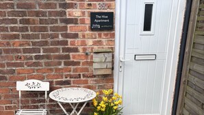 A brick wall with a door, a small table with a chair, and a potted plant with yellow flowers. - The Hive Apartment (Stockport)