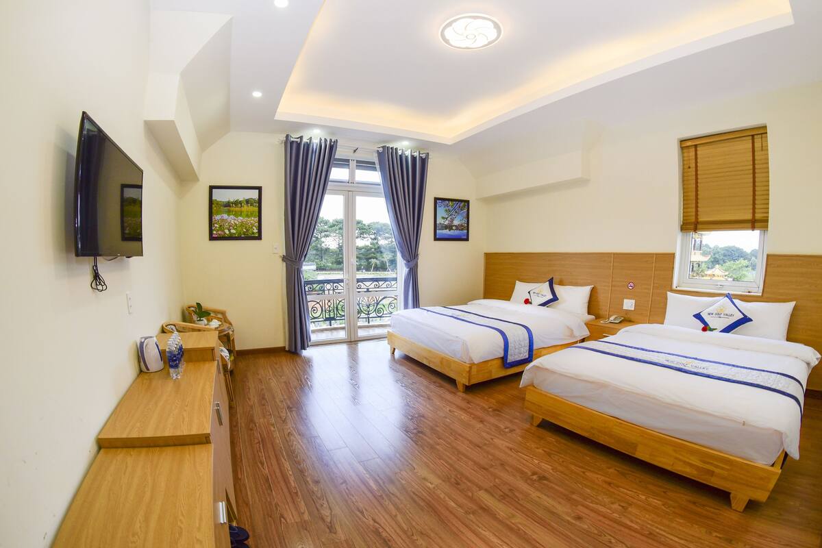 Quarto de hotel com duas camas, televisão, mesa com cadeira, janela com vista e varanda.