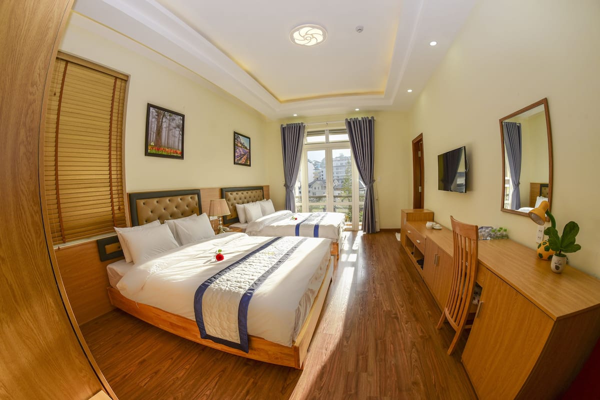 Quarto de hotel com duas camas, soalho de madeira, uma escrivaninha, uma cadeira, televisão e janela com cortinas.