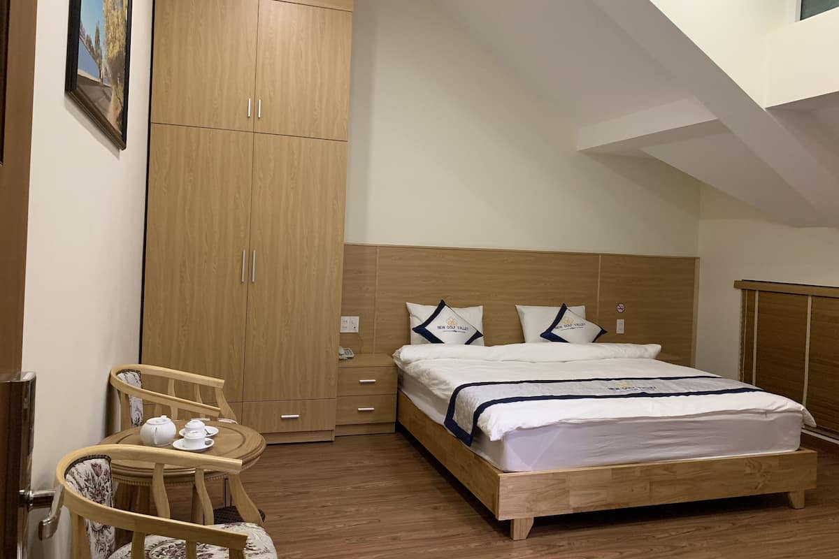 Quarto com cama de madeira, guarda-roupa de madeira, mesa redonda pequena com uma cadeira e um quadro na parede.