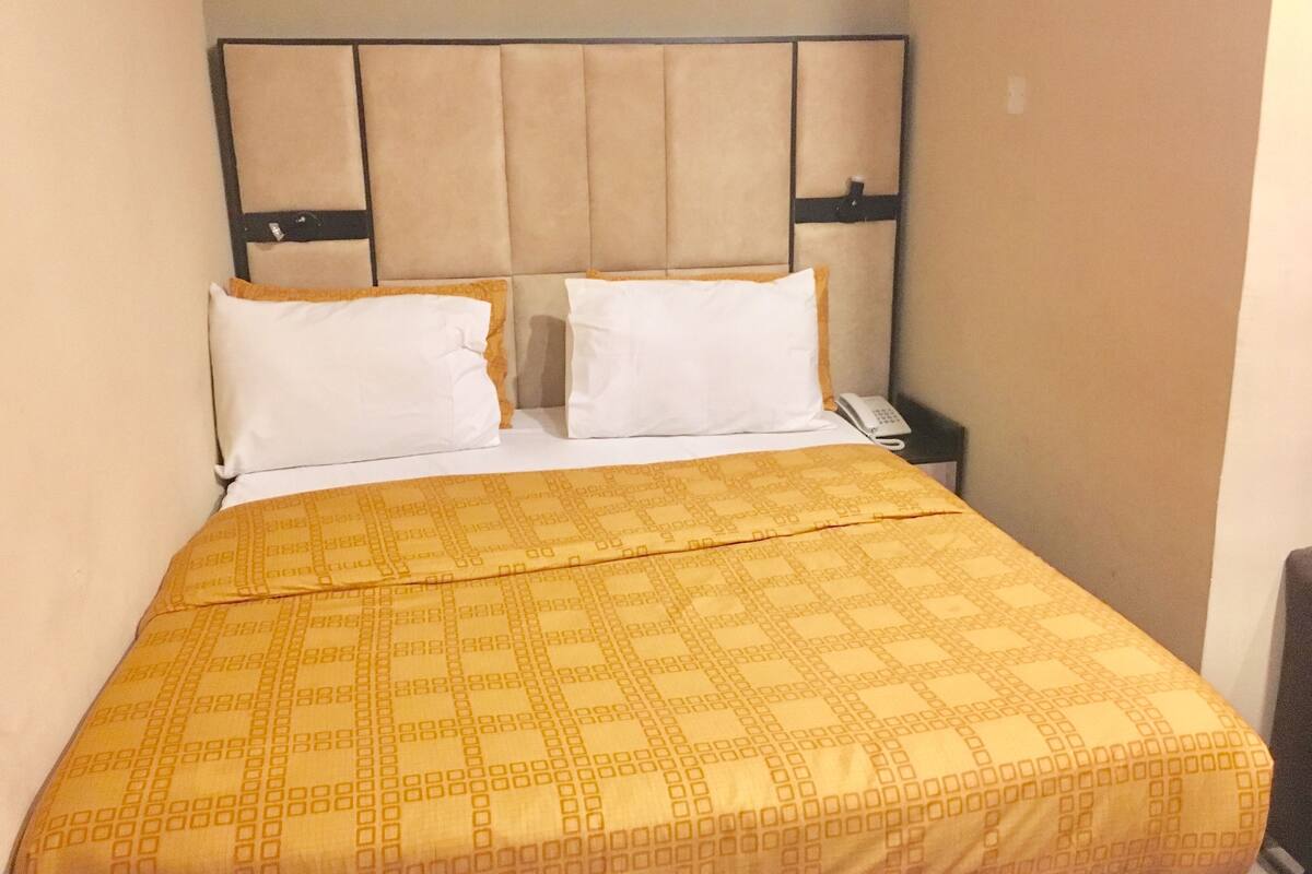 Une chambre d’hôtel avec un lit, deux oreillers blancs, une tête de lit beige et une couvre-lit à motifs.