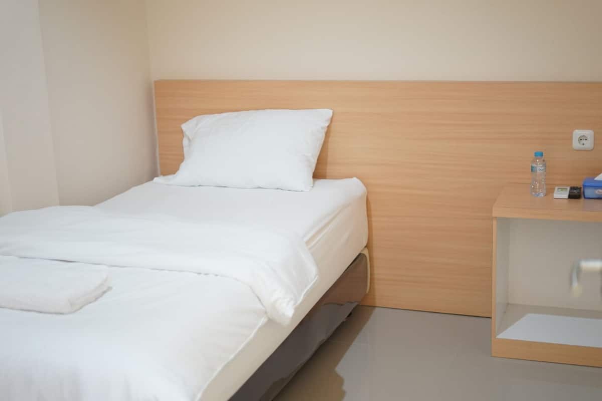 A single bed with white bedding, a wooden headboard, and a small bedside table with a water bottle and a box.