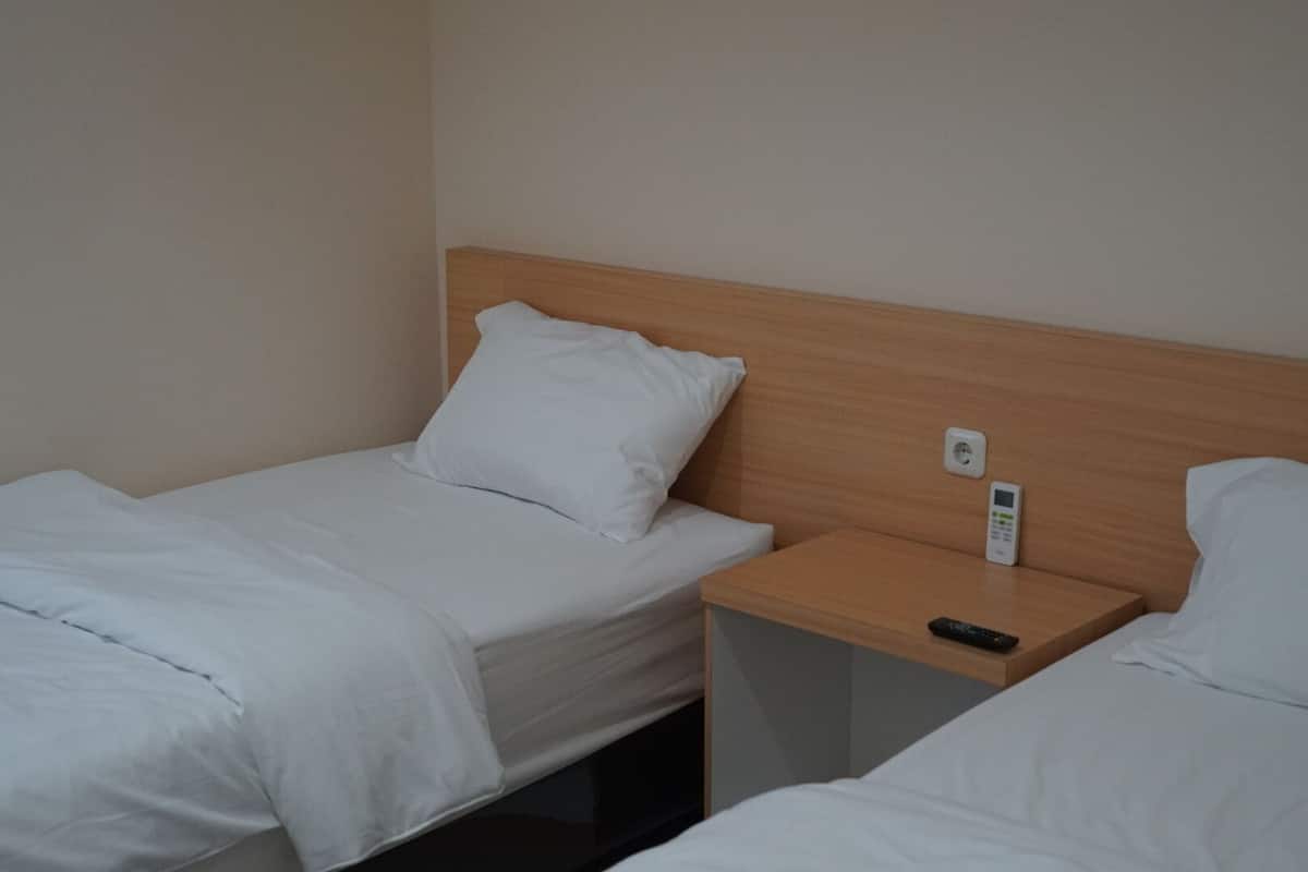 A twin bed with a wooden headboard, a bedside table with a remote control, and a power outlet on the wall.