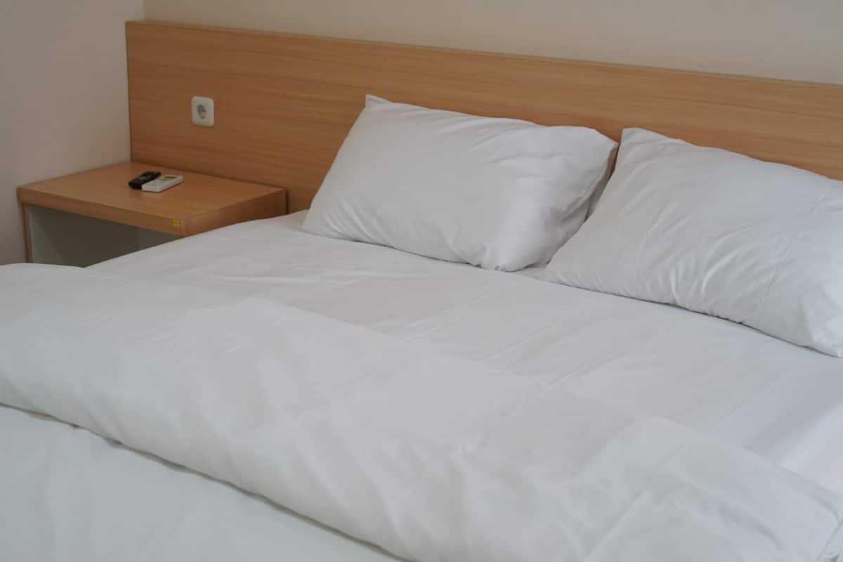 A neatly made bed with white linens and pillows, a wooden headboard, and a small wooden nightstand with a remote control.