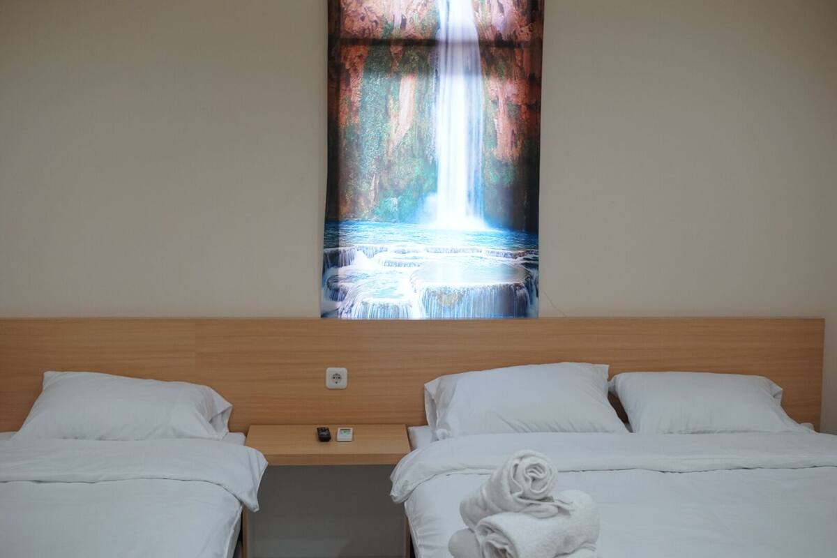 A hotel room with two beds, a wooden headboard, a wall-mounted painting of a waterfall, and a folded towel on the bed.