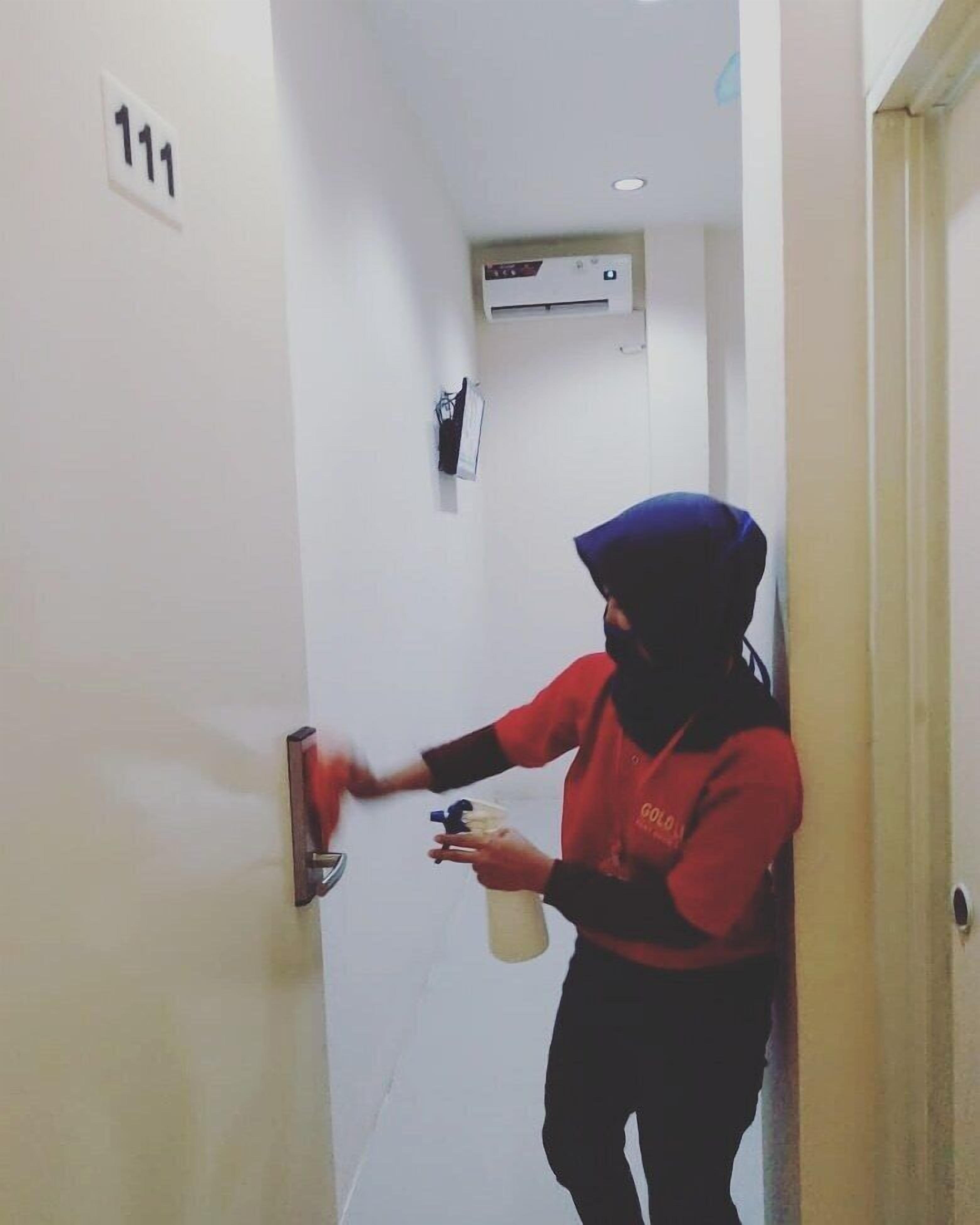 A person in a red uniform cleaning a door handle in a hallway with a number sign "111" on the wall and an air conditioning unit above.