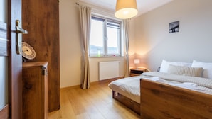 A bedroom with a wooden bed, a nightstand, a window with curtains, a radiator, and a wall clock. - Apartament Cichy - Apartamenty 5d (Swieradow-Zdroj)