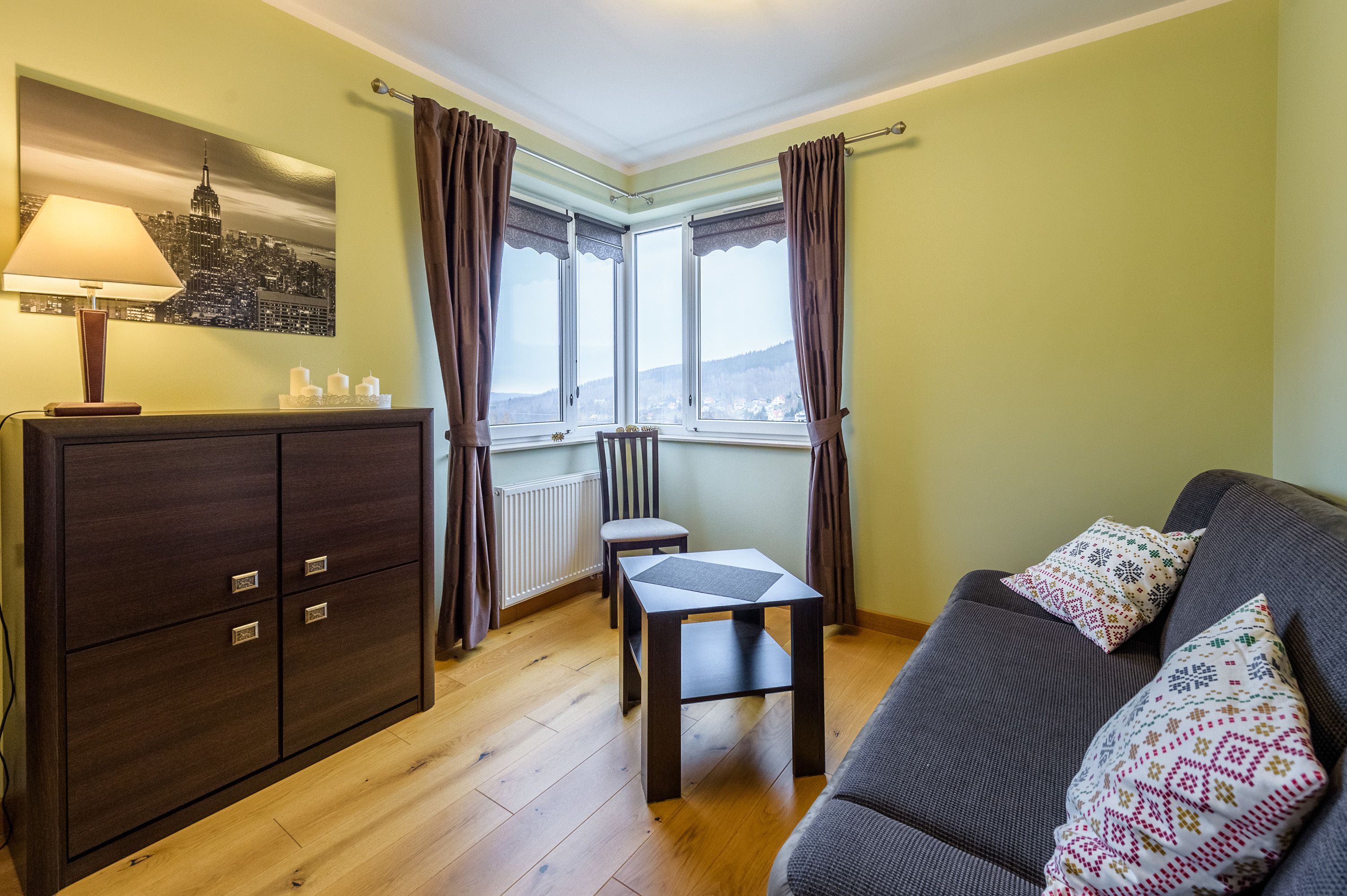 A cozy room with a dark wooden cabinet, a small table, a gray sofa with patterned cushions, a window with curtains, and a cityscape painting on the wall.