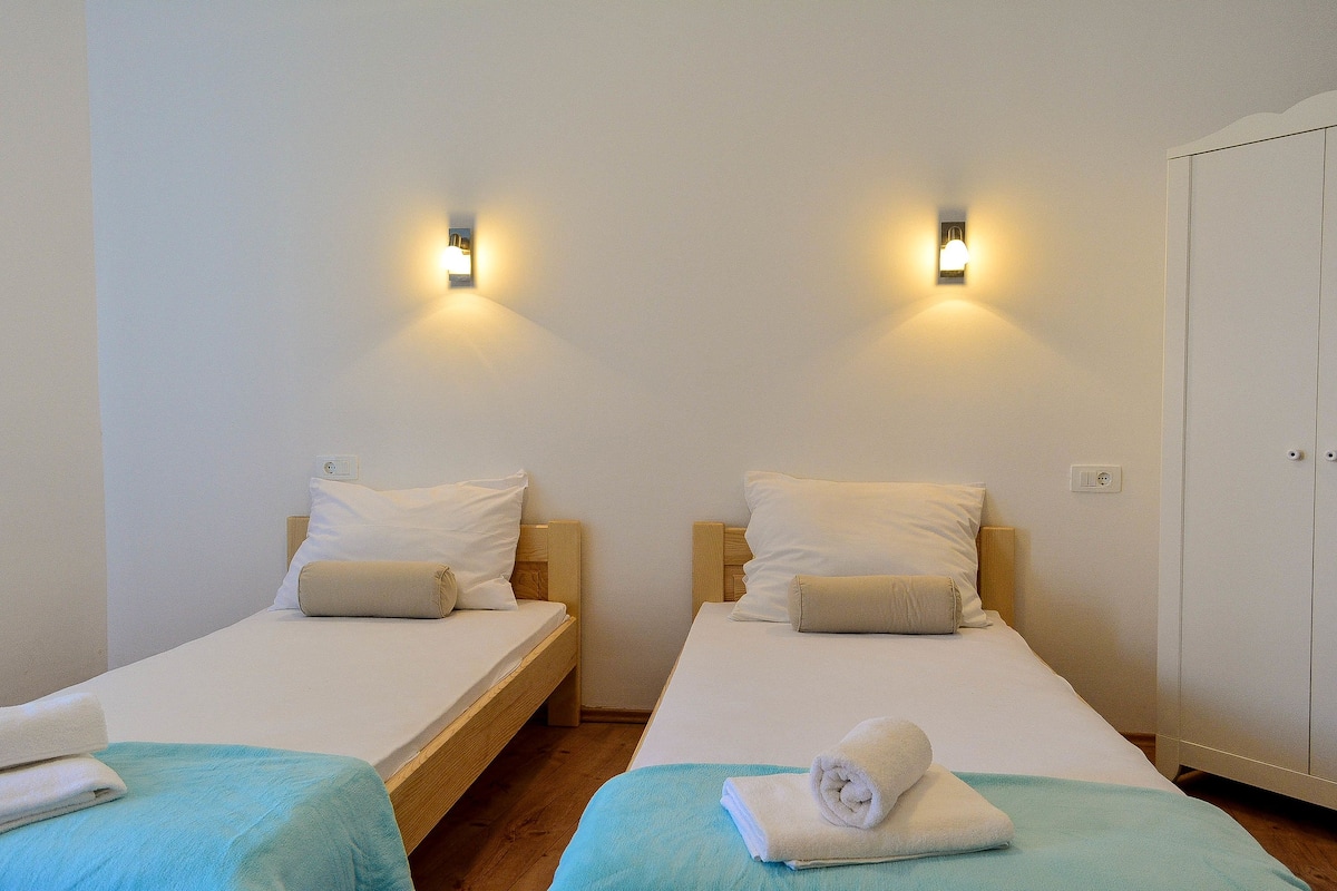 Une chambre avec deux lits simples, chacun doté d’une tête de lit en bois, de draps blancs et d’un plaid bleu. Des appliques murales sont fixées au-dessus de chaque lit.