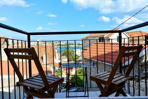 Balcón con dos sillas de madera y una mesa de metal, con vista a un pueblo costero de edificios con tejados rojos y mar azul.