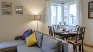 Een eetgedeelte met een ronde tafel voor vier personen, een grijze bank met kleurrijke kussens en een vloerlamp.