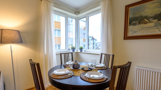 Een eetkamer met een ronde tafel voor vier personen, een raam met gordijnen, een radiator en een lamp.