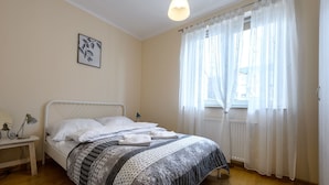 Een netjes opgemaakt bed met een bedsprei met patroon, een nachtkastje met een lamp en een raam met dunne gordijnen.