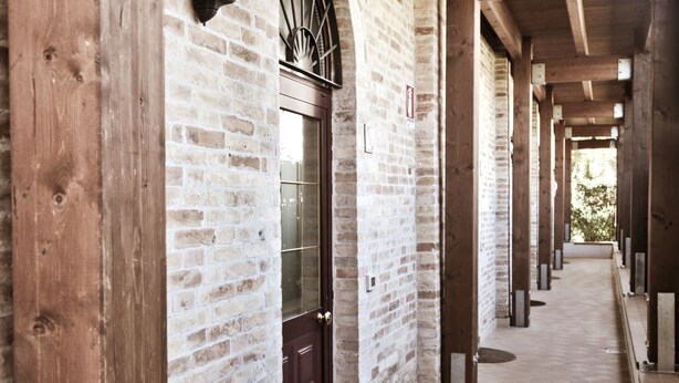 Un couloir couvert aux murs de briques, avec des poutres en bois et une porte dotée d’un panneau de verre.