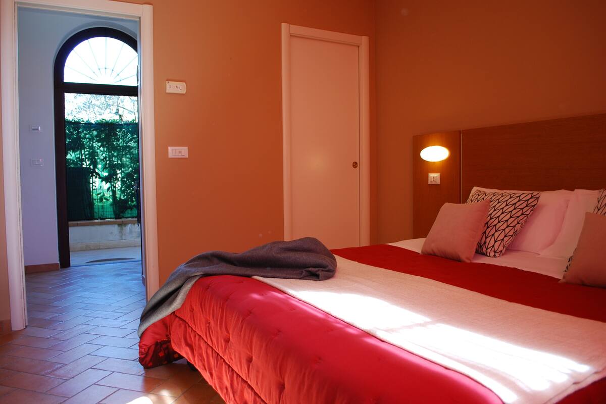 Habitación de hotel con cama roja, almohadas blancas, una puerta y una lámpara fijada a la pared.