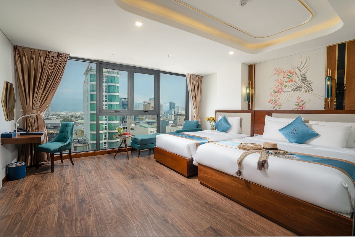 A hotel room with two beds, a desk, a chair, and a large window offering a city view.