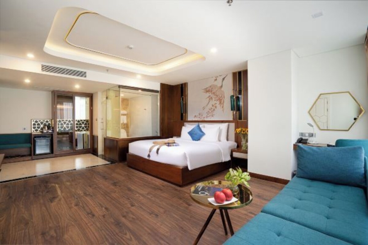 A modern hotel room with a large bed, a blue sofa, a glass coffee table with fruits, and a decorative wall piece.