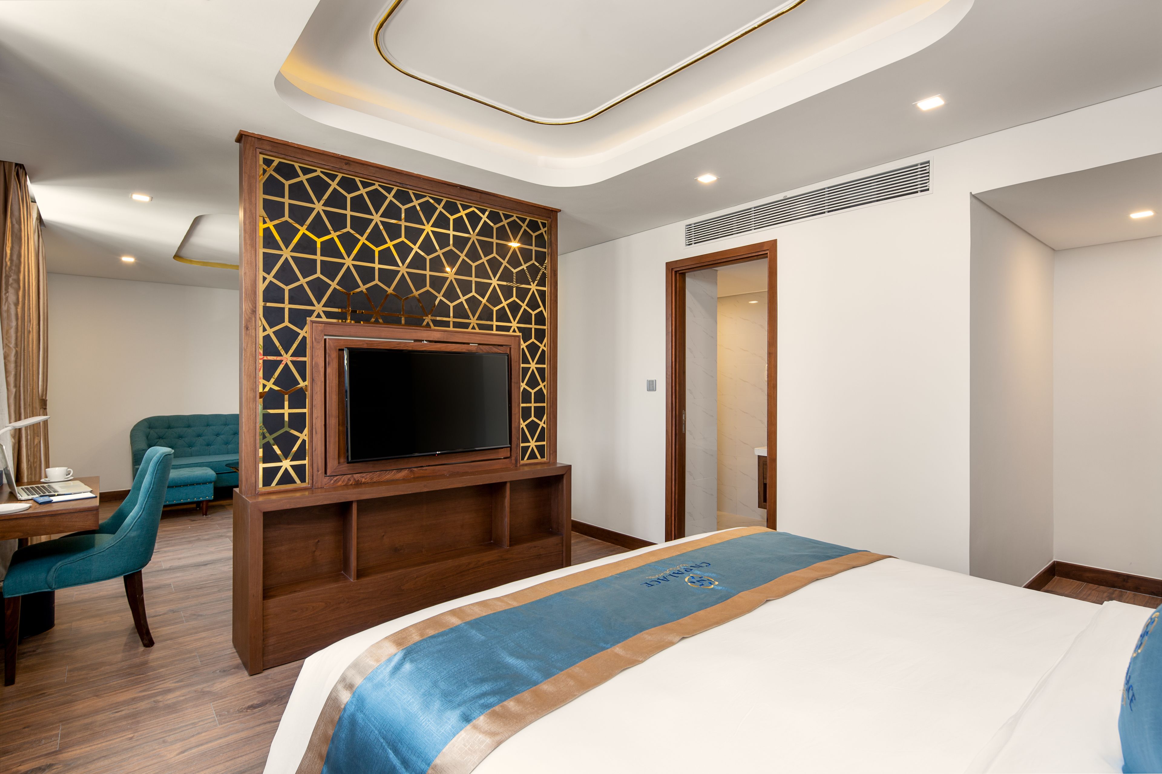 A hotel room with a large bed, a TV mounted on a wooden panel, a desk with a chair, and a door leading to another room.