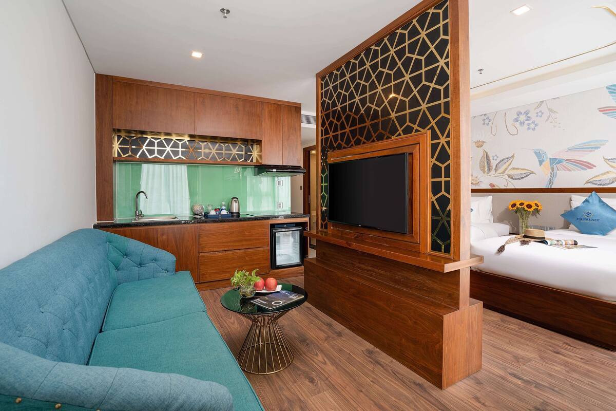 A compact living space with a sofa, a small table with a plant, a TV mounted on a wooden partition, and a kitchenette area.