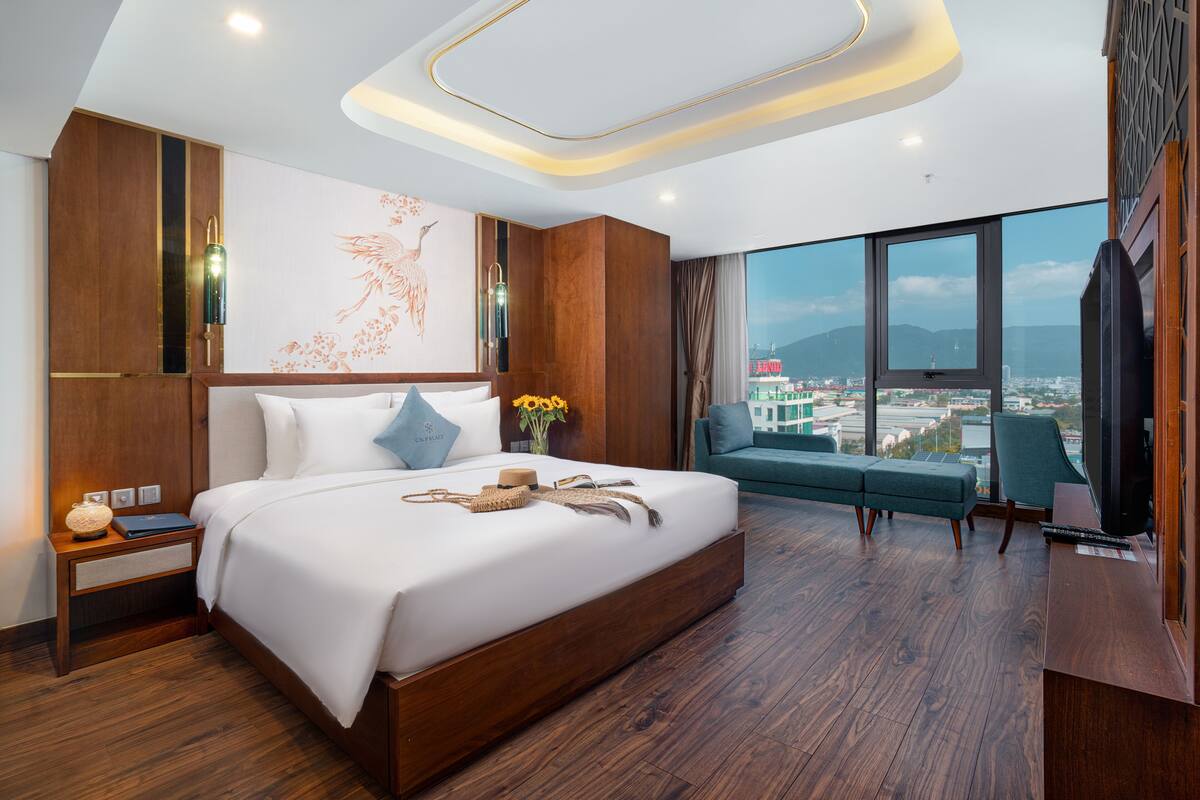 A modern hotel room with a large bed, a sofa, a TV, and a city view.