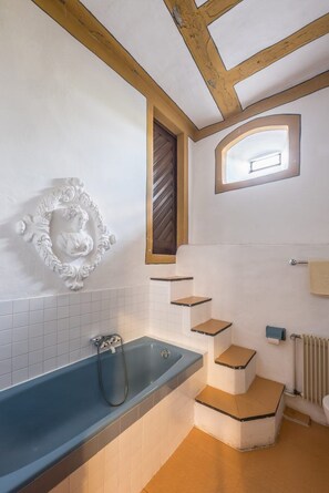 Ein Badezimmer mit einer Badewanne, Holzbalkendecke, weißer Wand mit dekorativer Reliefgestaltung und einem Fenster.