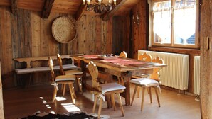A rustic wooden dining room with a large table, chairs, a chandelier, and a window with blinds. - Chalet Fiore (Val di Zoldo)