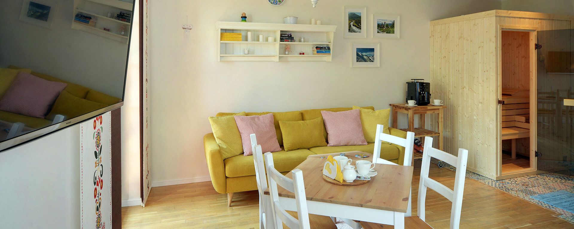 Un salon chaleureux avec un canapé jaune, des fauteuils blancs, une table basse en bois et une machine à café posée sur une table d’appoint.