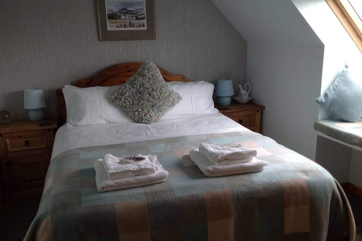 A neatly made bed with a checkered blanket, two pillows, and folded towels on a wooden nightstand.