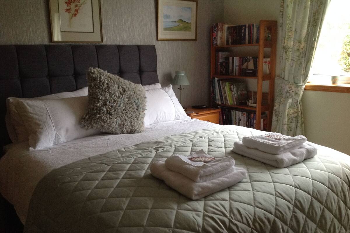 A neatly made bed with a quilted blanket and folded towels, a headboard, a bedside table with a lamp, and a bookshelf with books.