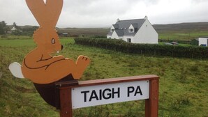 A sign with a rabbit silhouette and the text "TAIGH PA" in front of a white house with a grey roof, surrounded by green fields. - Hazelbank (Uig)
