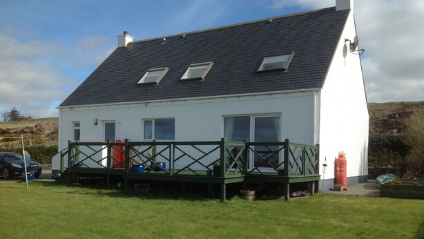 A white house with a dark roof, a green deck, and a red gas cylinder. - Hazelbank (Uig)