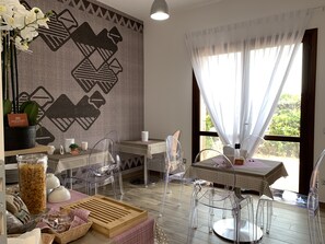 A modern room with a geometric patterned wall, a dining area with clear chairs and a table, and a large window with sheer curtains. - Relais del Borgo - Domo (Loiri Porto San Paolo)