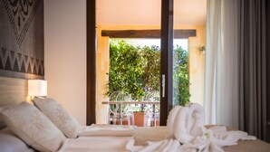 Habitación de hotel con cama, almohadas, manta y vistas a un balcón con sillas y una planta.