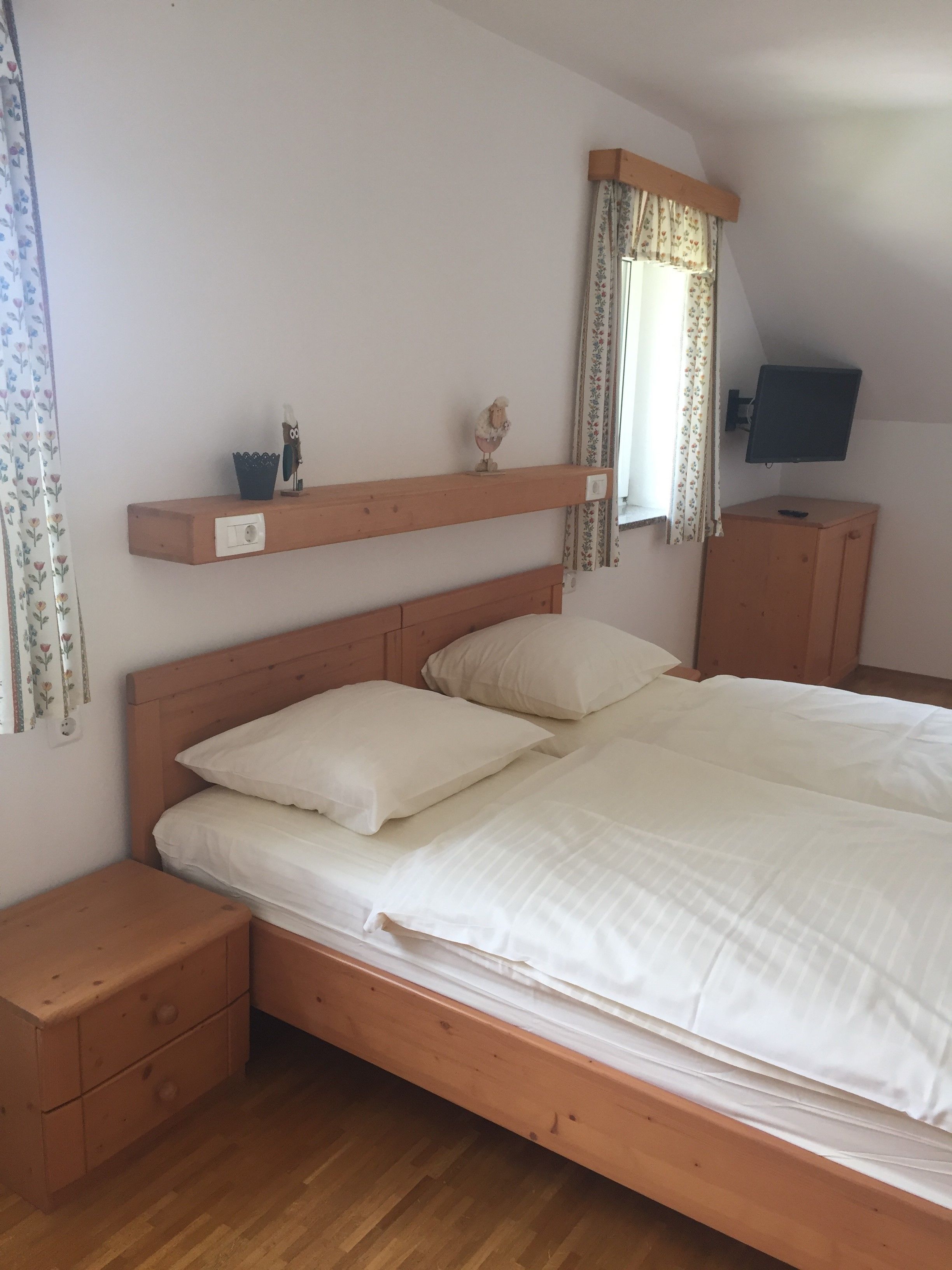 A bedroom with a wooden bed, two nightstands, a dresser, a mounted TV, and a window with floral curtains.