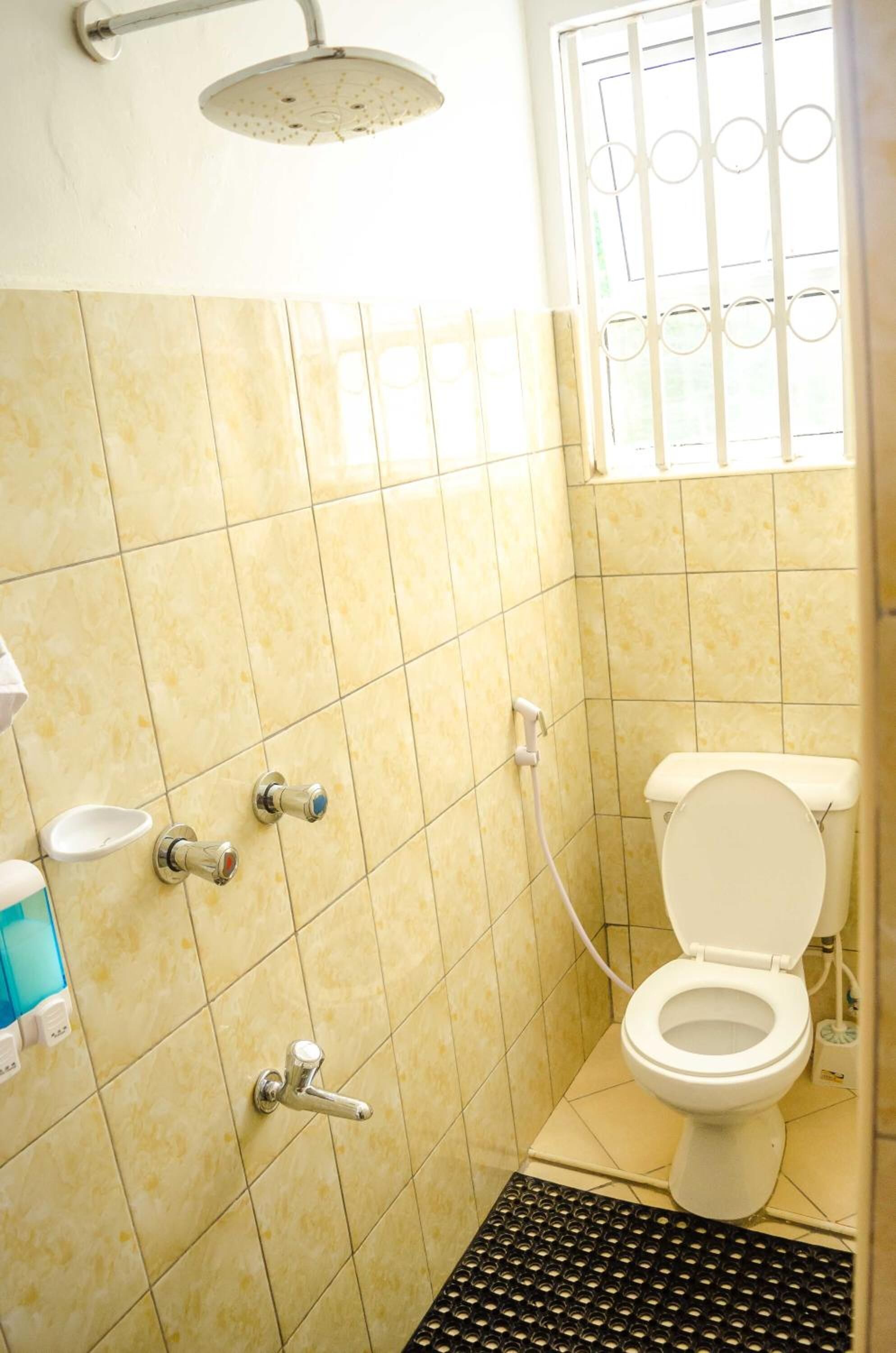 Un baño pequeño con azulejos amarillos, un inodoro blanco, un cabezal de ducha y una ventana con barrotes.