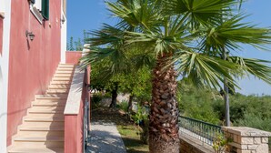 A red building with a staircase leading to a balcony, a palm tree, and a stone pathway. - Villa San Andreas (Zakynthos)
